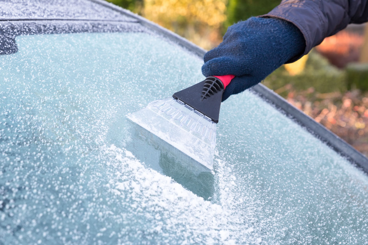 Hand wearing blue glove scratching ice from car window. The ice scraper is an extension of the arm. Part of the windshield is ice free. Only part of the window is visible. Symbol or concept of winter season, cold, frost, weather, sight.